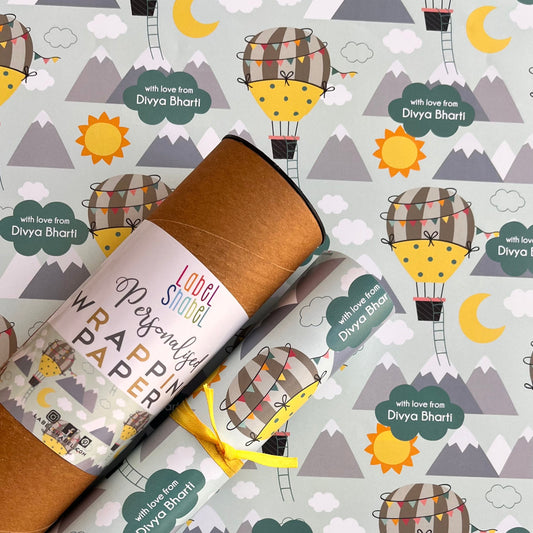 A roll of wrapping paper with a whimsical hot air balloon pattern, shown partially unrolled over a matching patterned surface. The wrapping paper features a light gray background with repeating illustrations of yellow crescent moons, brown and yellow hot air balloons, gray mountains, and green labels reading 'with love Divya Bharti'.