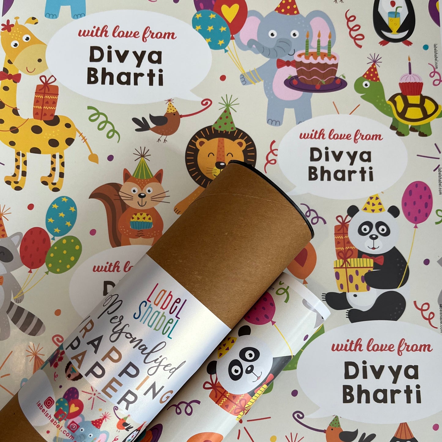 A personalized cardboard tube gift wrapper rolled up and tied with a red ribbon, placed on top of colorful birthday-themed wrapping paper printed with cartoon animals and the text 'with love from Divya Bharti'.