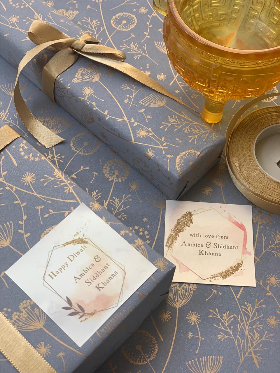 A beautifully wrapped gift box for Diwali, wrapped in dusty blue paper with a gold floral pattern, tied with a gold ribbon. The box has a white square label that reads 'Happy Diwali Ambika & Siddhant Khanna' in gold foil.
