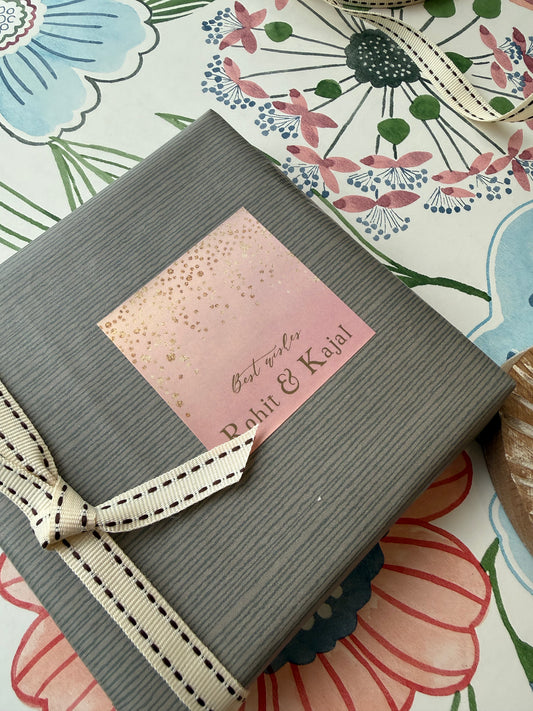 A gray ribbed gift box tied with a cream ribbon, featuring a pink square label that reads 'Best wishes Rohit & Kajal' in gold foil, placed on a floral-patterned surface.