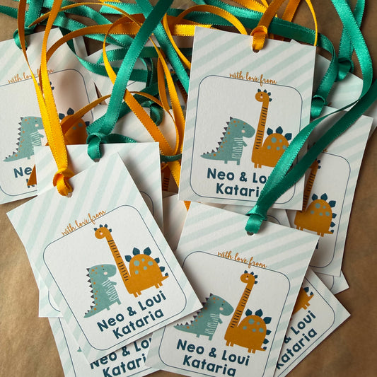 A pile of personalized dinosaur-themed gift tags with ribbon loops, featuring cartoon dinosaurs and the names 'Neo & Loui Kataria' on a light background.