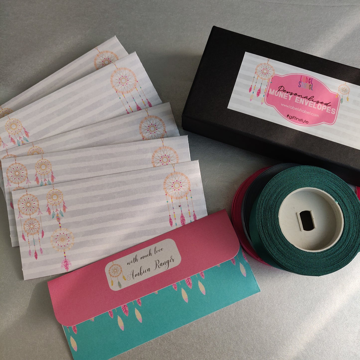 A gift set featuring a black box labeled 'Dreamcatcher Money Envelopes' with a pink label, a roll of dark green ribbon, and several white envelopes printed with dreamcatcher designs, arranged on a gray surface.