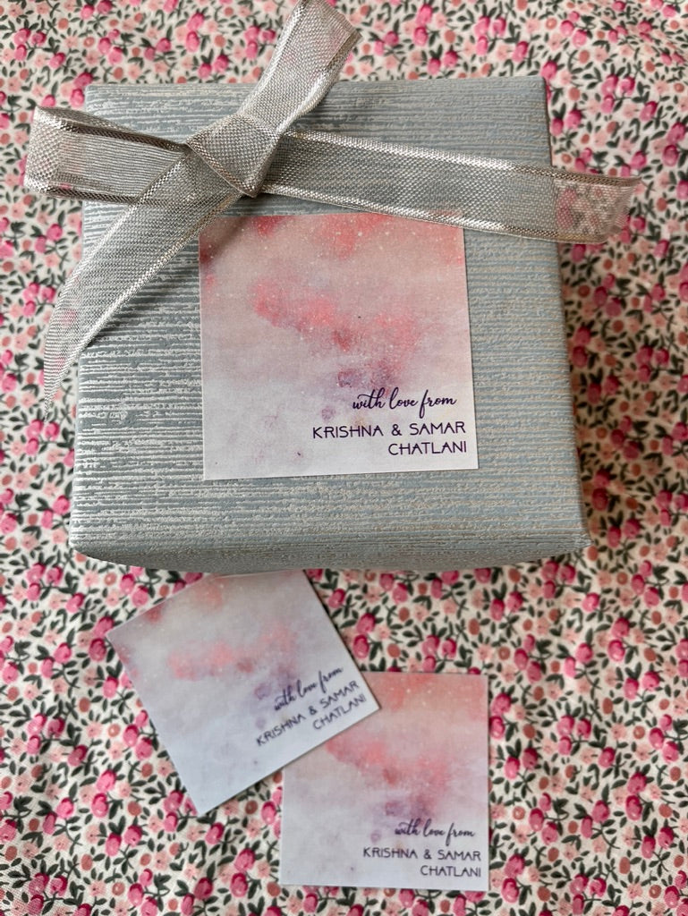 A silver gift box wrapped with a sheer silver ribbon and a watercolor-style label reading 'with love from KRISHNA & SAVAR CHATLANI,' placed on a pink and black floral fabric background with two matching cards below.
