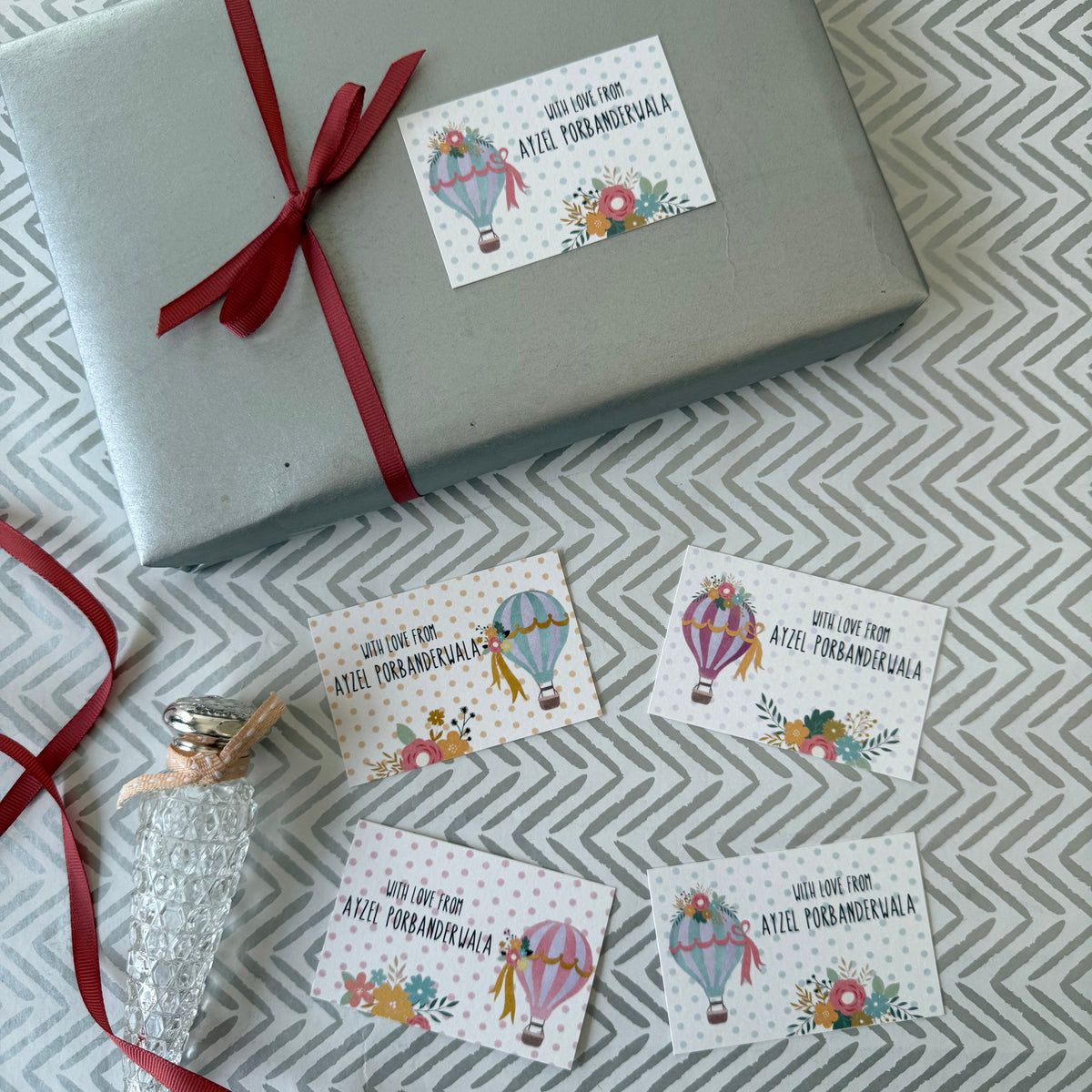A wrapped gift box in light gray with a red ribbon and several small printed gift tags featuring hot air balloon illustrations and the text 'Will one day give to you Ate Pambankula' are arranged on a gray-and-white chevron fabric background.