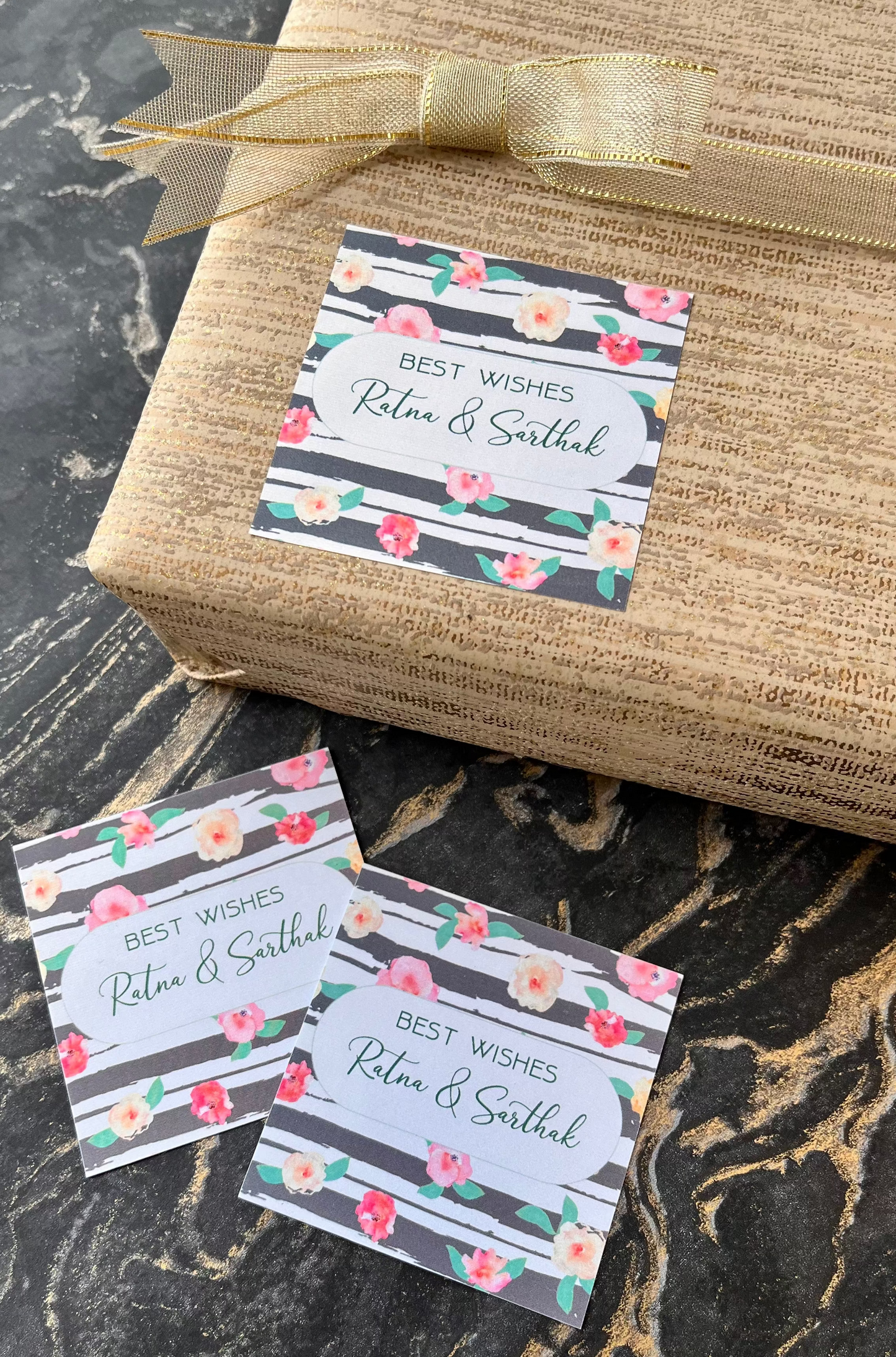 Three floral gift tags with gray striped borders and the text 'BEST WISHES Ritu & Sarthak' are arranged on a burlap-wrapped gift box tied with a gold ribbon, set on a dark marble surface.