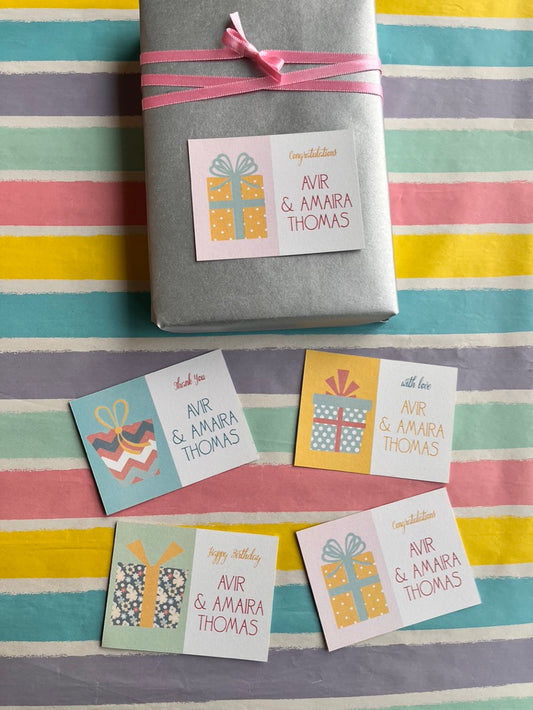 A wrapped gift box tied with a pink ribbon sits on a pastel striped surface, with four small gift tag cards arranged below. The tags have birthday greetings