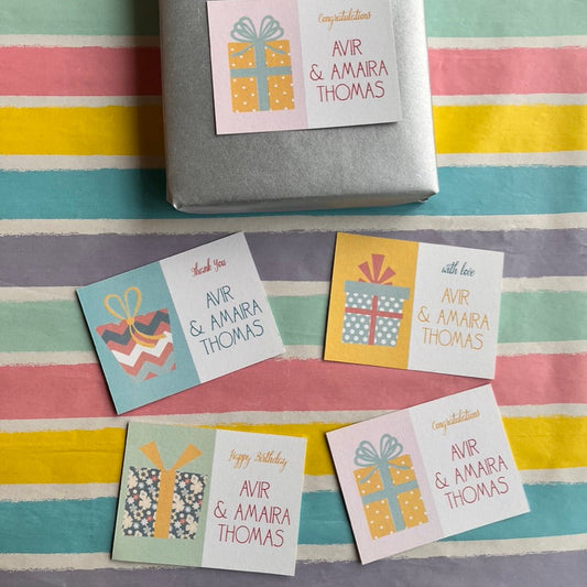 A wrapped gift box tied with a pink ribbon sits on a pastel striped surface, with four small gift tag cards arranged below. The tags have birthday greetings