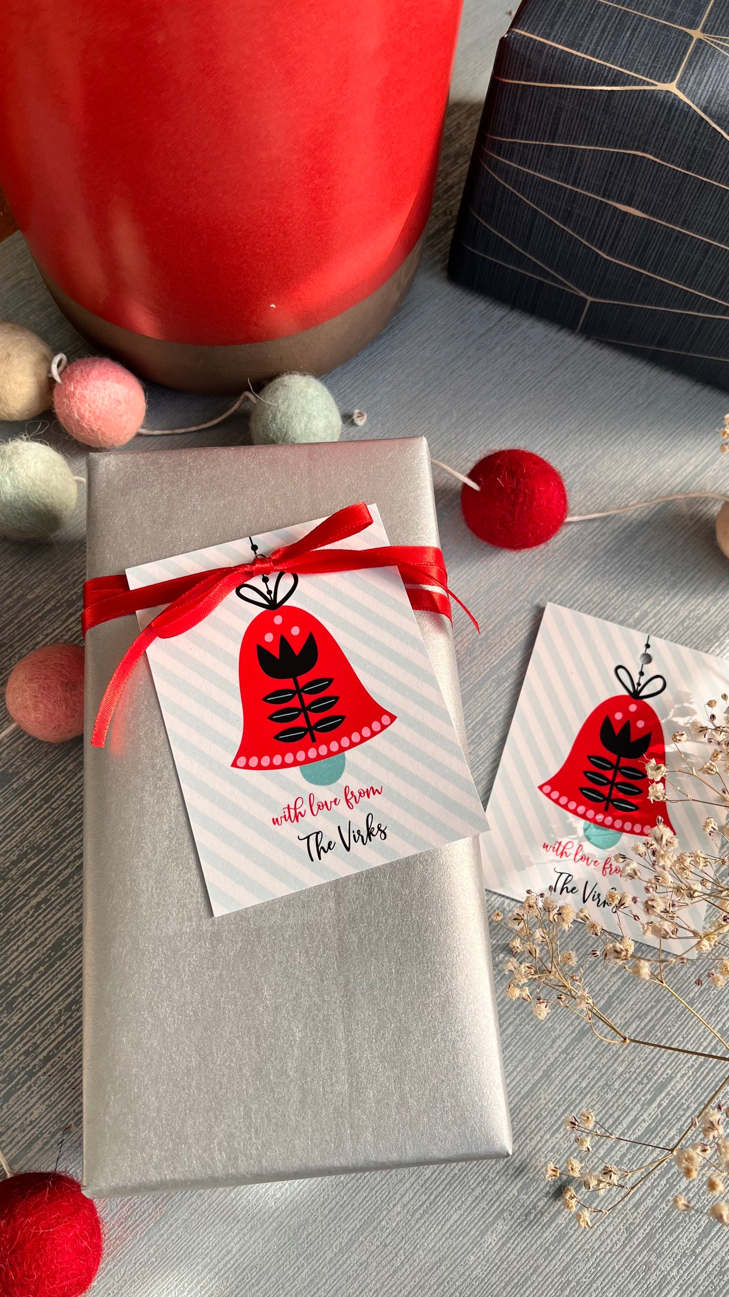 Ribbon tags with 'Holiday Bells' design, attached to a gift wrapped in a red ribbon, presented on a table with decoration items and a cup in the background.
