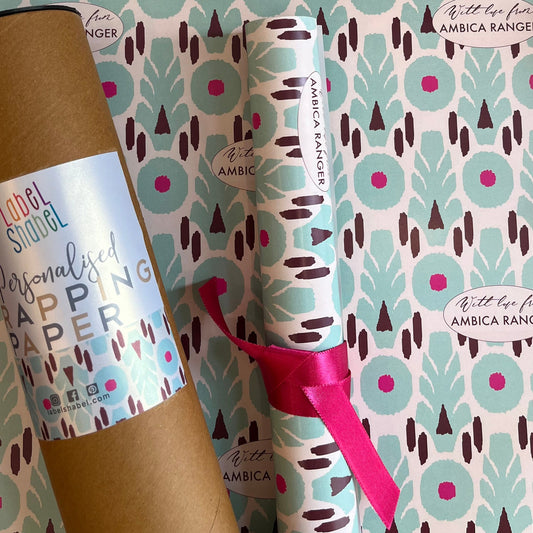 Personalized wrapping paper set by Amrica Ranger, featuring a kraft paper tube and a rolled sheet of the patterned paper tied with a pink ribbon, displayed against the patterned paper background.