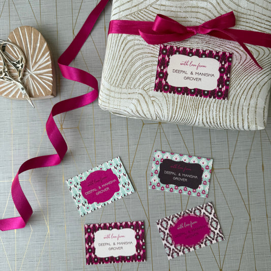 A wrapped gift box tied with a bright pink ribbon and a matching gift tag, arranged on a light gray patterned surface with several sample gift tags and a small wooden heart-shaped box nearby.