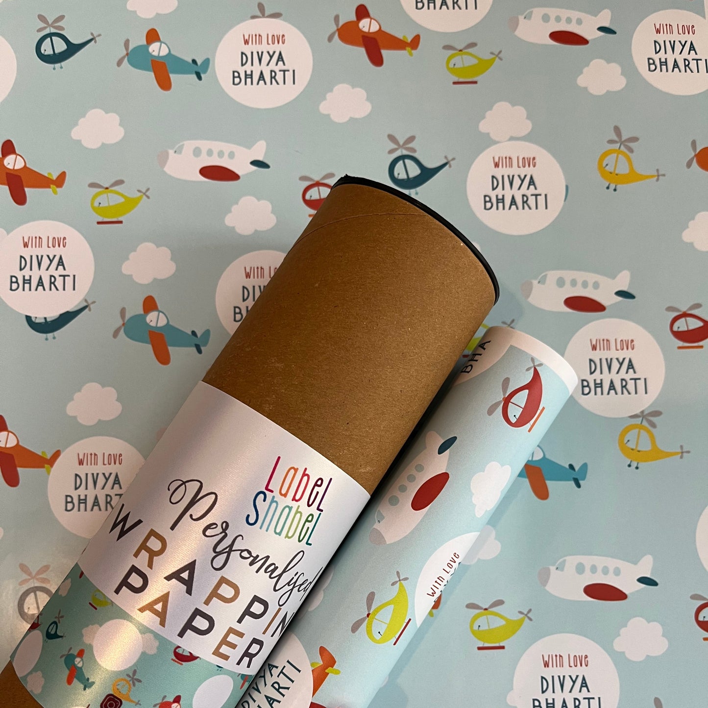 A roll of wrapping paper placed on a light blue patterned background with cartoon planes helicopters and clouds