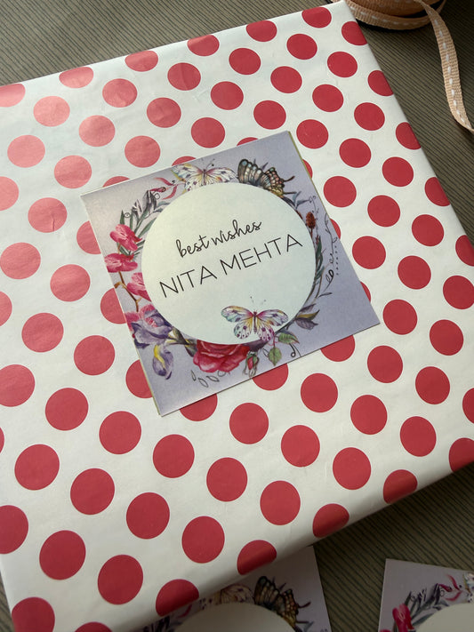 A square gift box wrapped in white paper with large red polka dots, featuring a central floral label in lavender hues with flowers