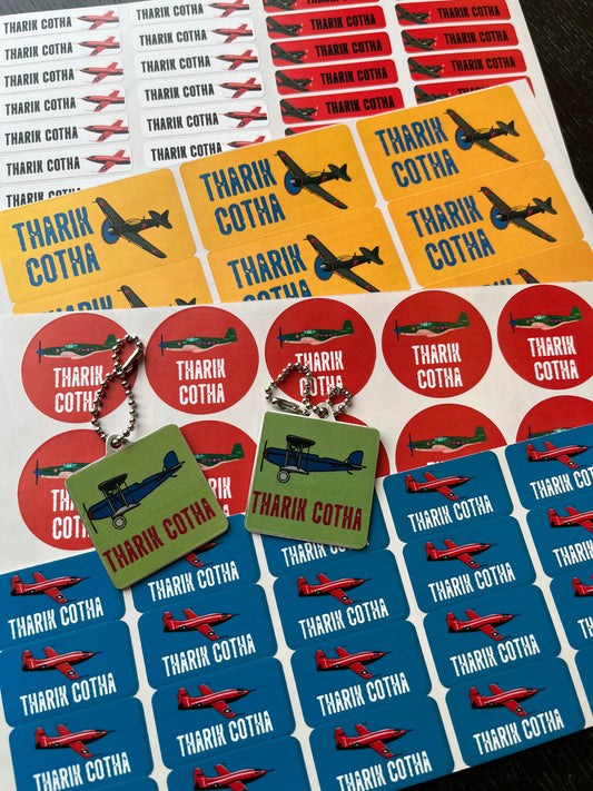 A sheet of colorful sticker sheets featuring a name with red, yellow, blue, and green airplane illustrations. Two small square keychains with matching stickers are displayed in front on a dark surface.