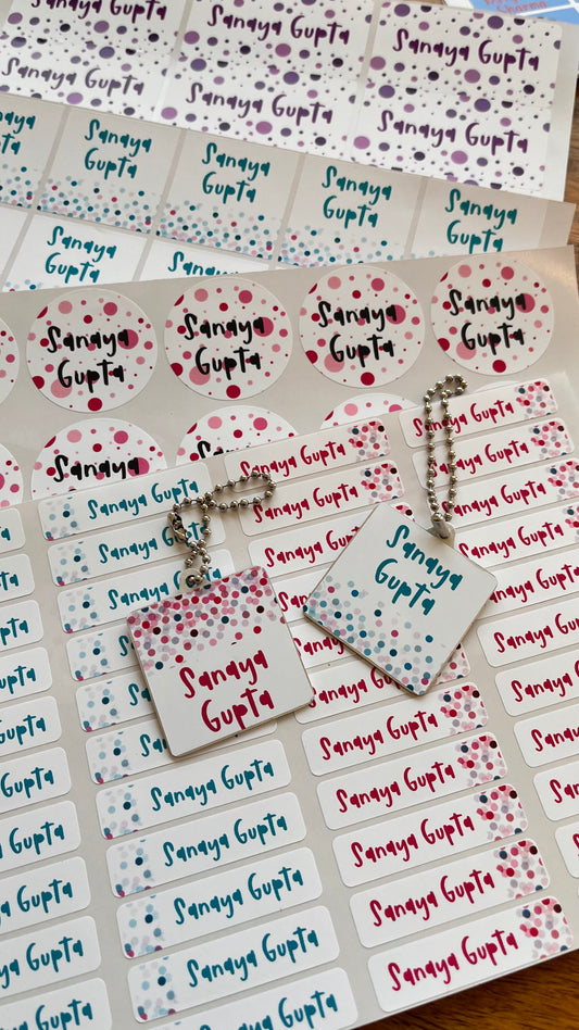 Custom printed name labels and matching circular stickers featuring the name 'Sanaya Gupta' in blue and red script with colorful confetti dots, displayed on a wooden surface.