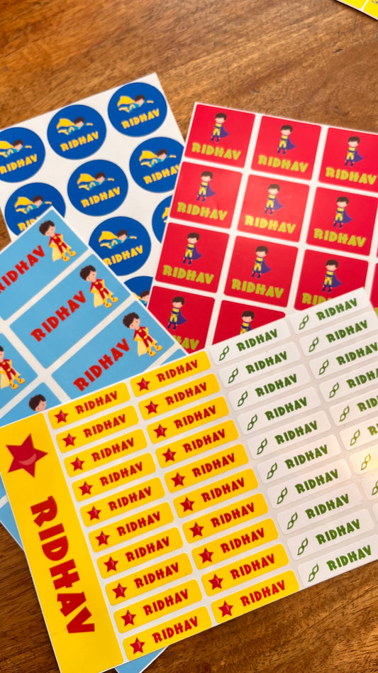 Several printed sticker sheets laid out on a wooden surface, featuring the name RIDHAv in multiple colorways including blue, red, yellow, and white all with superhero cartoon character