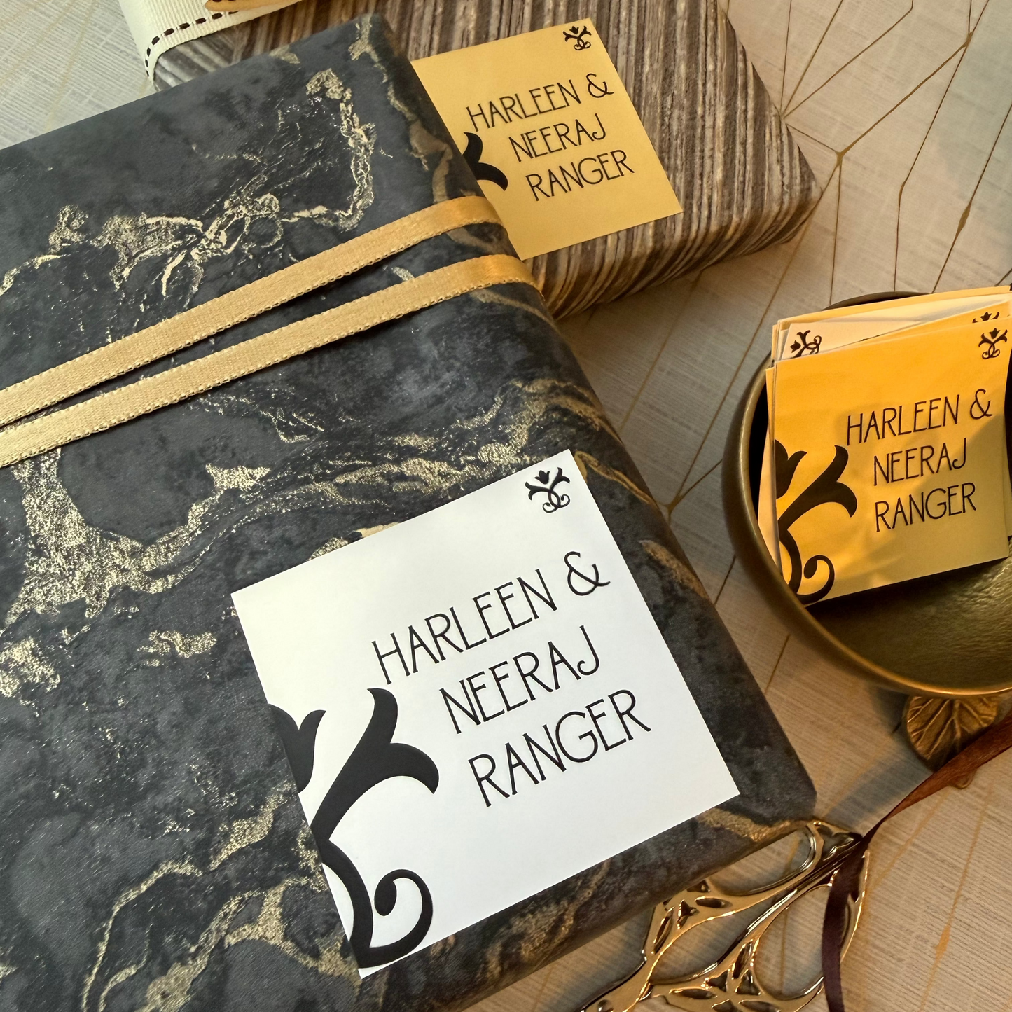 Three custom gift boxes wrapped in black and gold marble paper, a striped box with a cream bow, and a small gold tray holding matching yellow name tags, all labeled 'Harleen & Neeraj Ranger' on a light wood background.