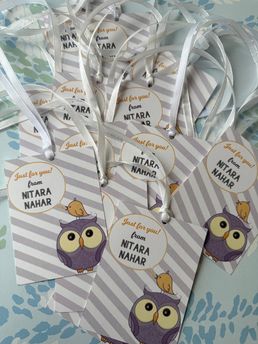 A pile of personalized gift tags printed with a purple owl and a small bird, with white ribbons attached, on a light blue patterned surface. The tags read 'Just for you! from NITARA NAHAR'.