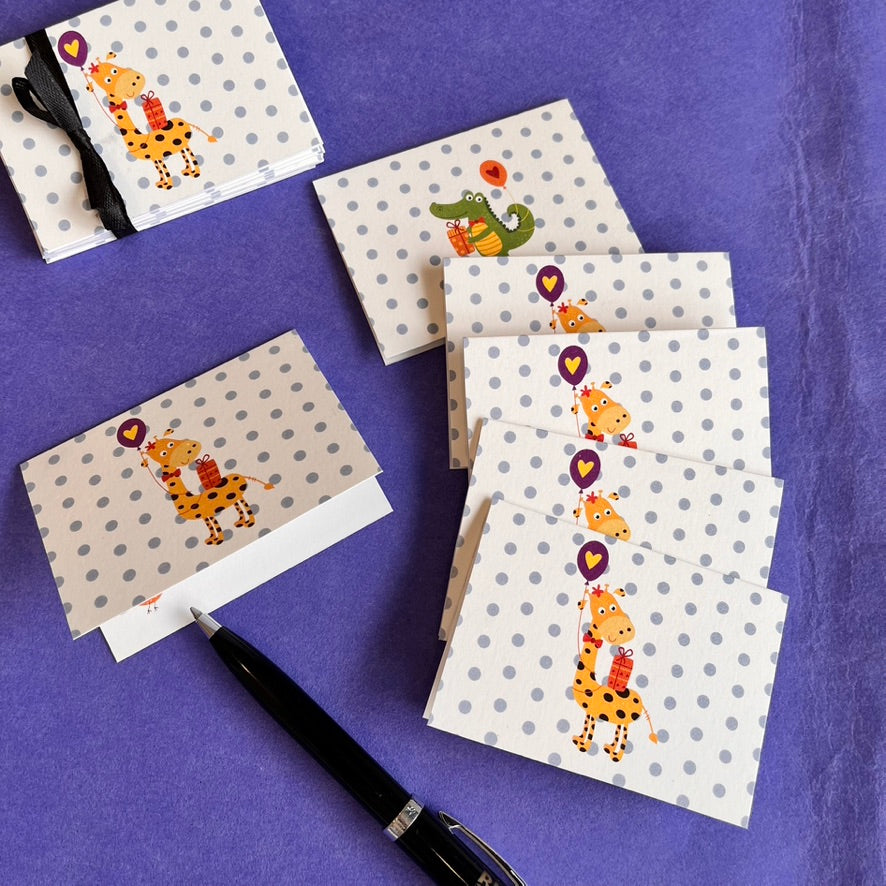 A set of children's greeting cards and matching envelopes with a cartoon giraffe holding a purple heart-shaped balloon, arranged on a textured purple fabric background next to a black pen.