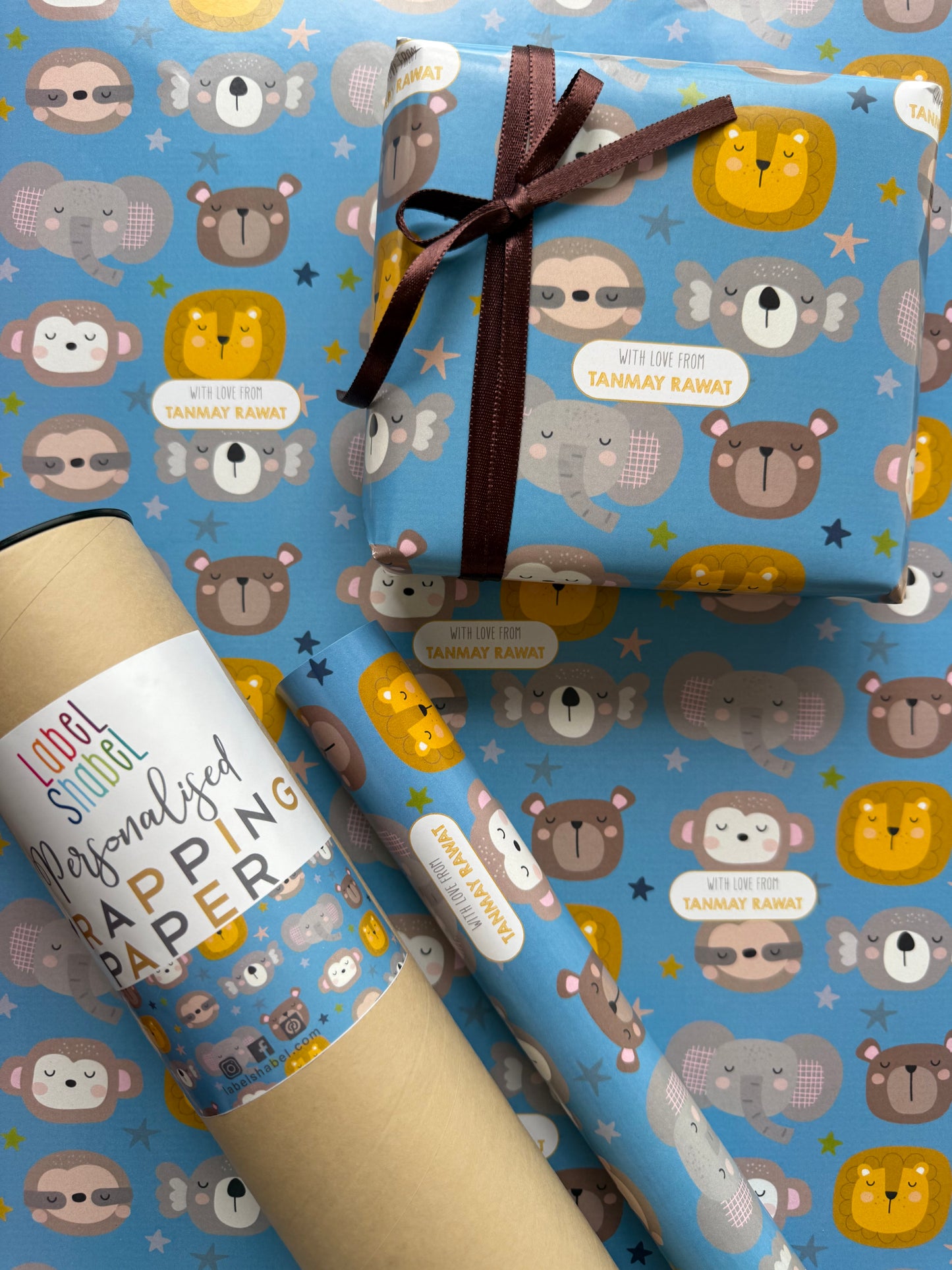 A roll of wrapping paper and a wrapped gift box with a matching pattern. The wrapping paper has a light blue background decorated with cartoon animals in yellow, brown, and gray, along with small white labels.