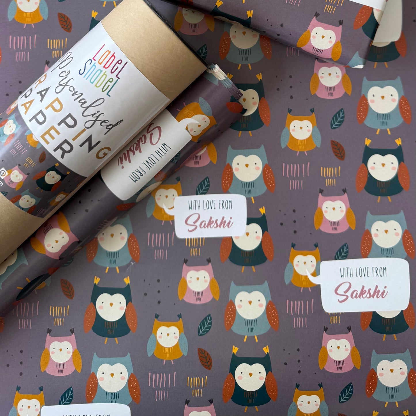 Owl-themed wrapping paper and gift wrap set with a repeating pattern of cute owls on a dark gray background, shown wrapped around gift boxes and a roll with a red ribbon.