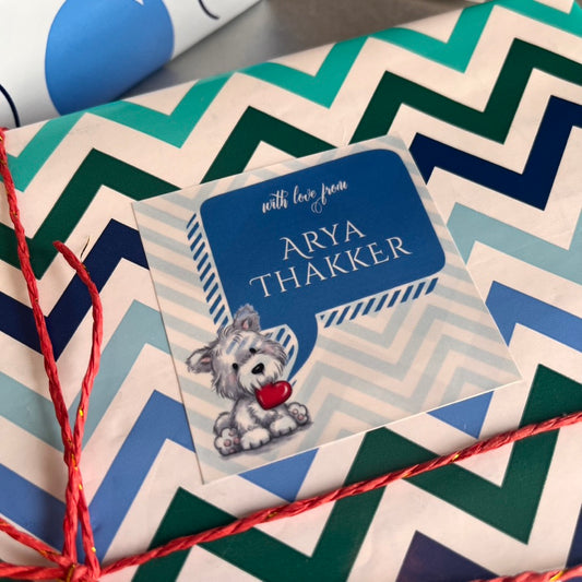 A close-up of a personalized gift tag with a blue speech-bubble design reading 'with love from ARYA THAKKER' and a small dog illustration, placed on top of chevron-patterned wrapping paper with red string.