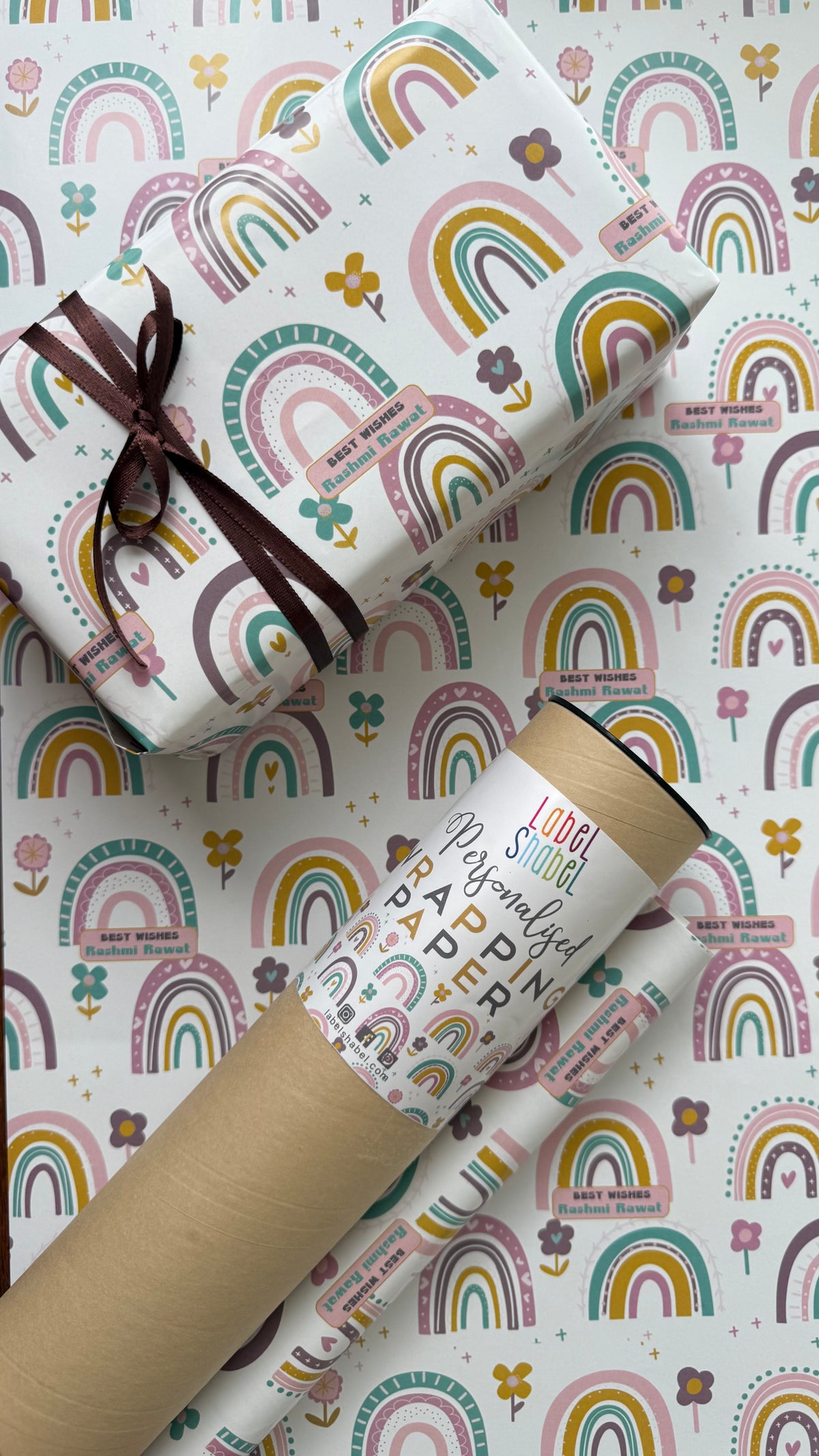 A roll of wrapping paper and a wrapped gift box featuring a whimsical pastel rainbow and flower pattern on a white background, tied with a dark brown ribbon.
