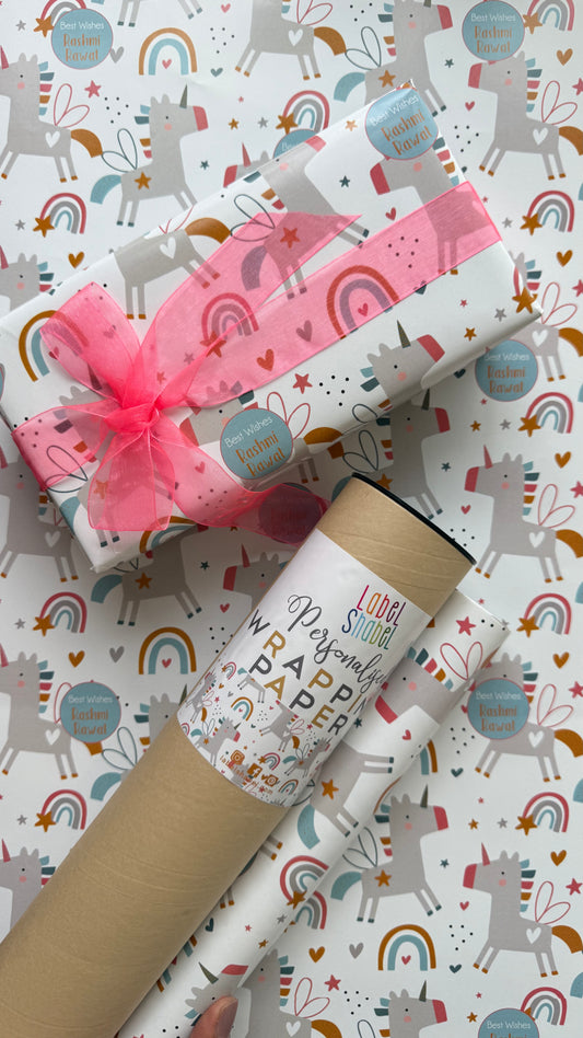 Unicorn and rainbow patterned wrapping paper shown wrapped around a gift box with a pink ribbon and a cardboard mailing tube, on top of a matching rolled sheet of the same paper.