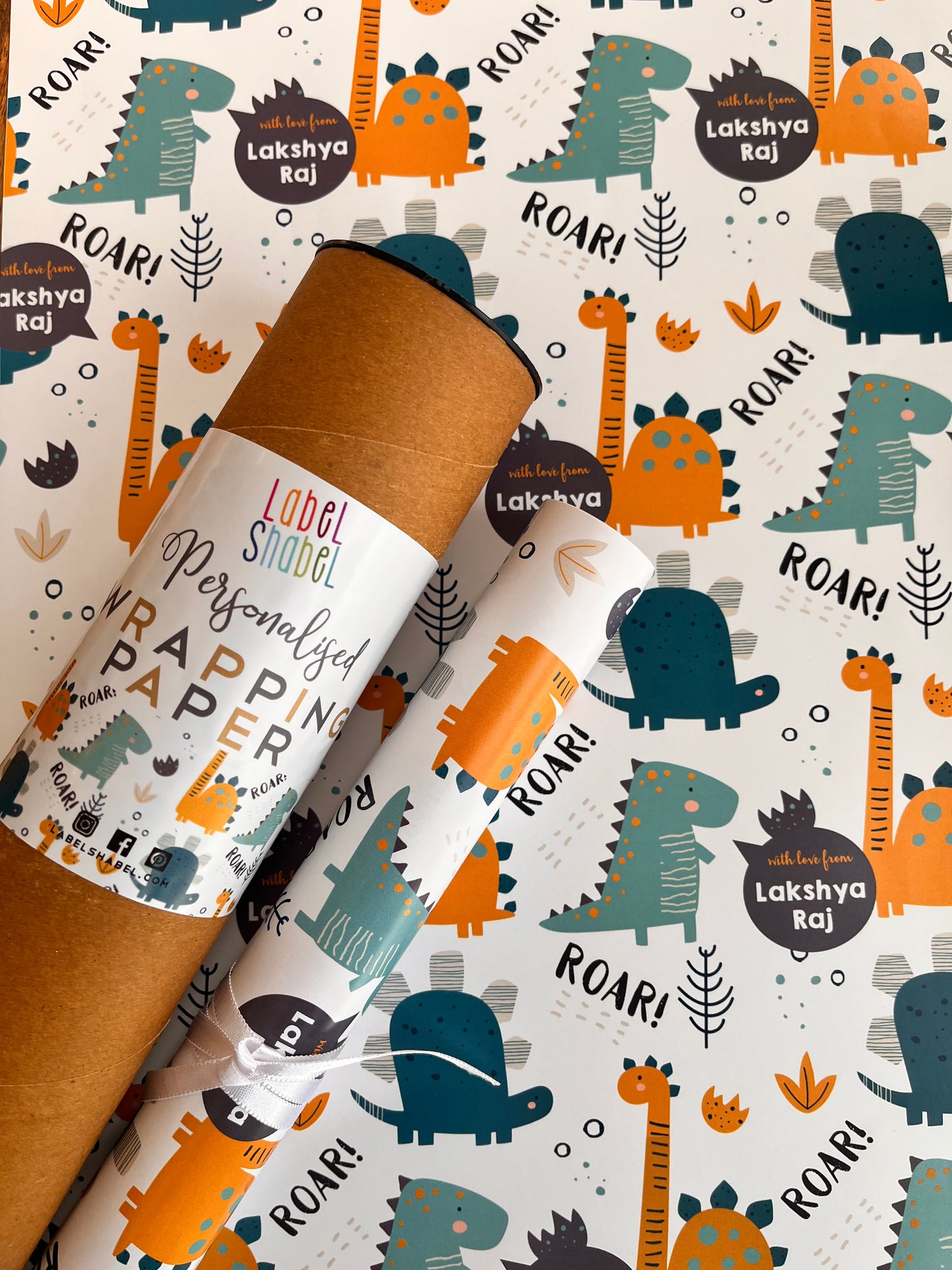 Two rolls of wrapping paper with a playful dinosaur and sound-themed pattern, displayed diagonally on top of a matching roll of brown kraft paper. The pattern features cartoon dinosaurs in teal and orange, with text like 'ROAR!'