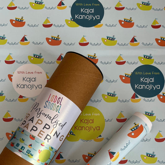 Personalized wrapping paper set featuring a brown kraft cardboard tube and rolled sheets printed with a colorful sailboat pattern, tied with an orange ribbon, on a matching patterned background. Visible text includes 'Kajal Kanojiya' and 'Love From Kajal Kanojiya'.