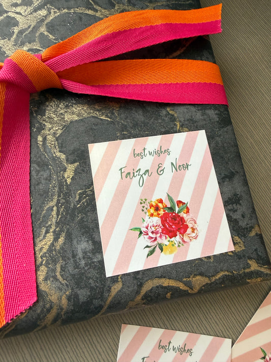 A close-up of a gift box wrapped in a dark gray and gold marbled paper, decorated with a bright pink, orange, and yellow striped ribbon. A square card with pink diagonal stripes, a floral illustration, and the text 'best wishes From & To' is tucked inside the box.
