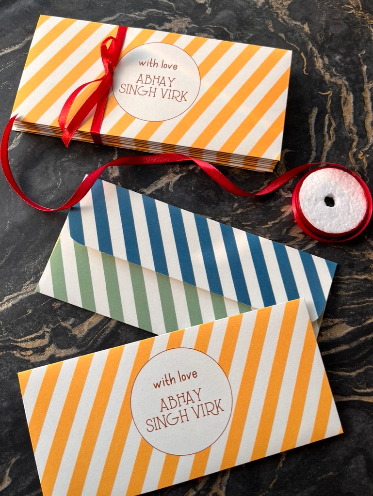 Three striped gift envelopes or cards arranged on a dark marble surface, with orange-and-white diagonal stripes on the top item and blue-and-white stripes on the middle item. A third envelope has green-and-white stripes.