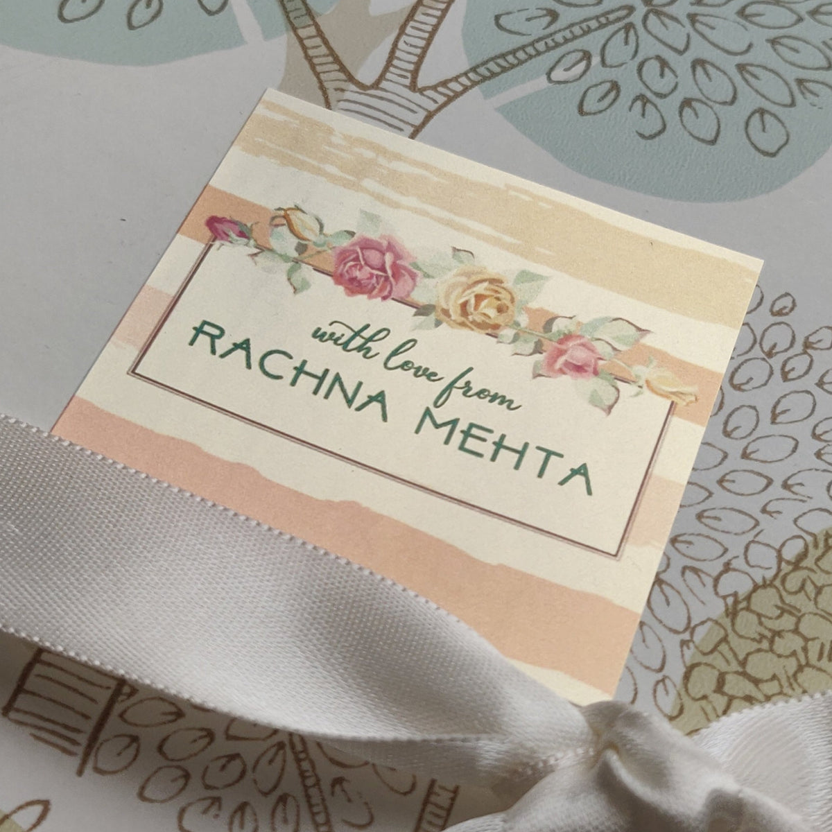 A close-up of a gift box wrapped in a patterned paper featuring dandelion and floral designs in muted green, beige, and white, tied with a white satin ribbon. A small card on the front reads 'with love, RACHNA MEHTA'.