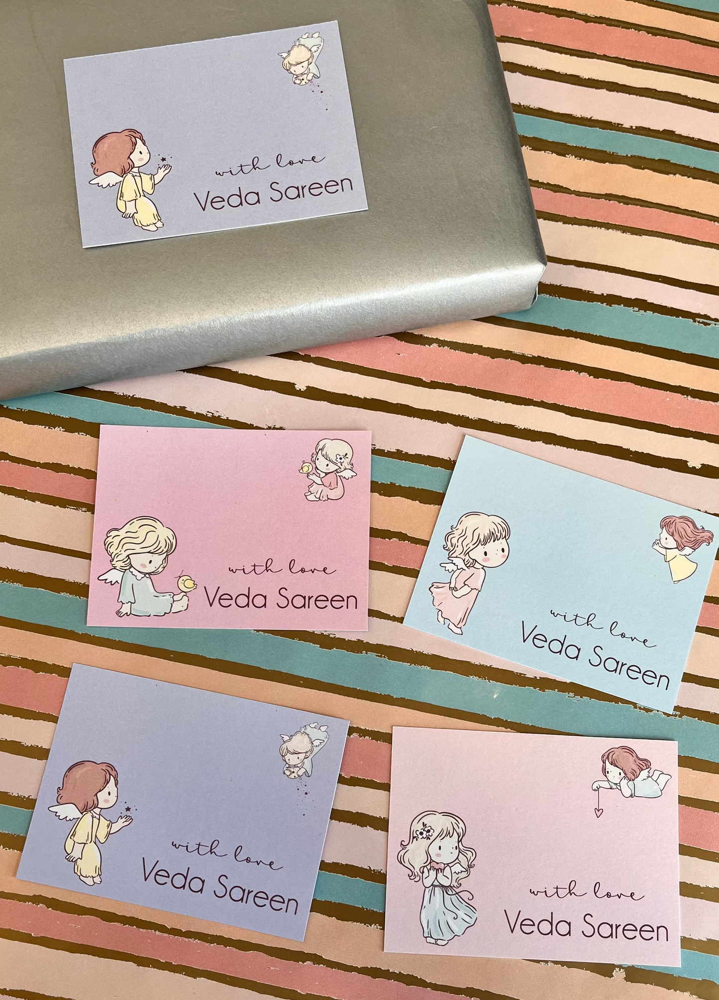 Four pastel greeting cards with cute anime-style girl illustrations and the text 'with love Veda Saren' are arranged on a striped pastel surface, with a silver box partially visible at the top.