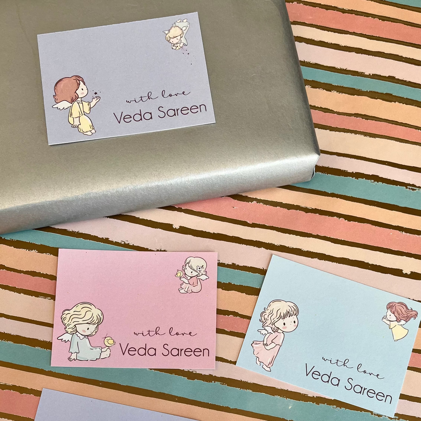 Four pastel greeting cards with cute anime-style girl illustrations and the text 'with love Veda Saren' are arranged on a striped pastel surface, with a silver box partially visible at the top.