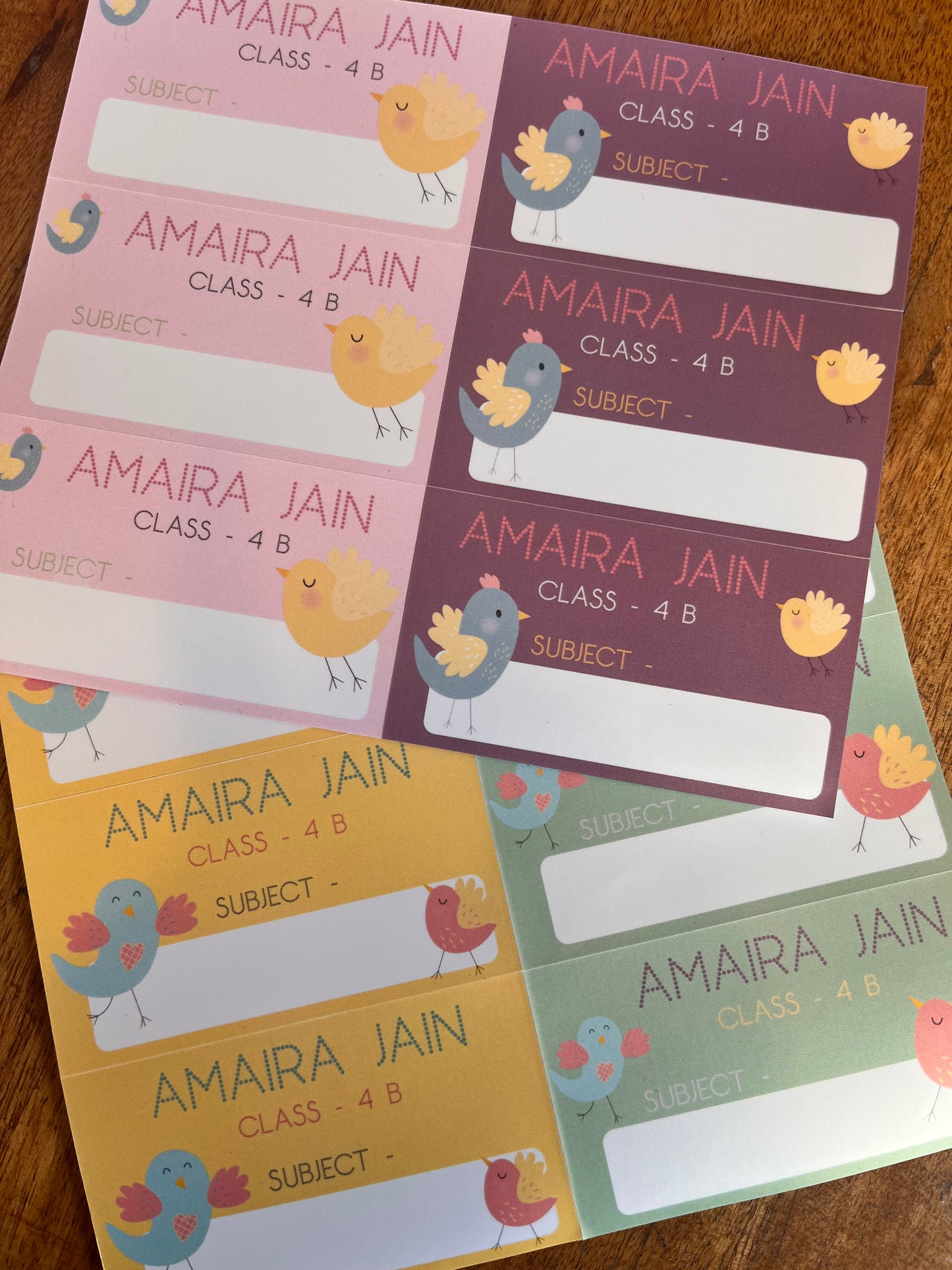colorful book labels with cartoon bird illustrations, arranged in a fan shape on a wooden surface. The labels feature different pastel colors (purple, pink, green, yellow)