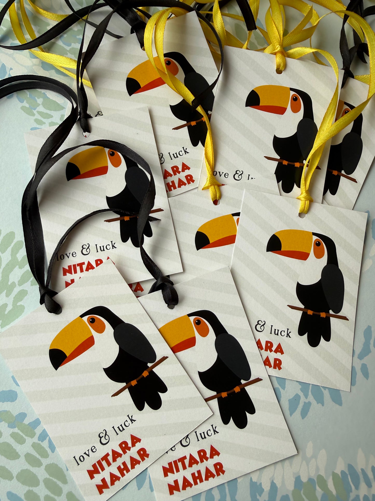 Several white gift tags with toucan illustrations and hanging ribbons are arranged on a light patterned surface. The tags include the text 'love & luck' and 'NITARA MAHAR'.