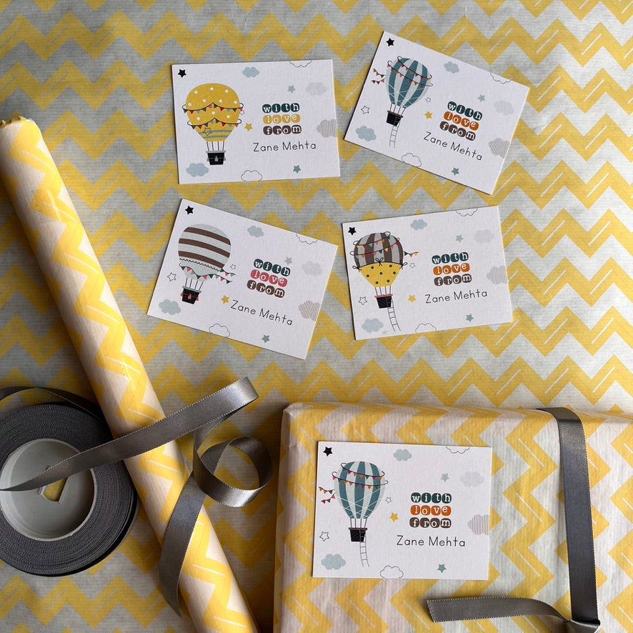 A styled flat-lay of hot air balloon themed wrapping paper and ribbon. The image shows four flat sheets of wrapping paper with different balloon designs and text reading 'Cute Hot Air Balloon from Zone Mehta', a roll of yellow and white chevron patterned wrapping paper, a roll of gray tape, and a roll of gray ribbon on a yellow and white chevron background.