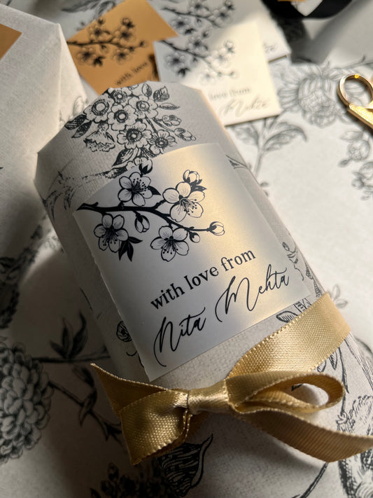 A close-up of a cylindrical gift box wrapped in white paper with black floral line art and a gold ribbon bow, placed on matching patterned wrapping paper.
