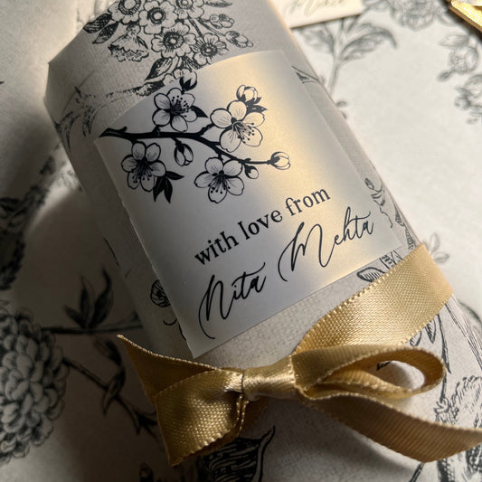 A close-up of a cylindrical gift box wrapped in white paper with black floral line art and a gold ribbon bow, placed on matching patterned wrapping paper.