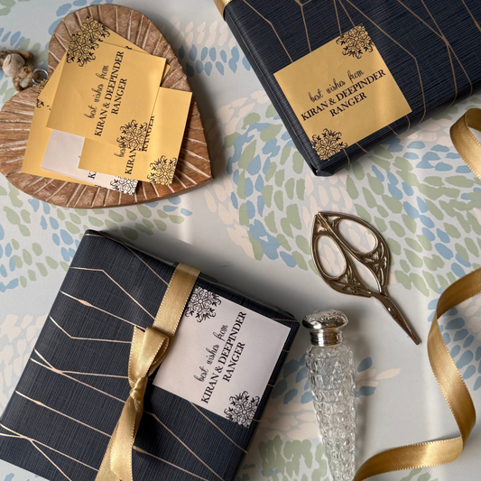 gift labels adorn navy wrapped gift boxes with gold geometric lines and gold ribbons are arranged on a floral-patterned surface.