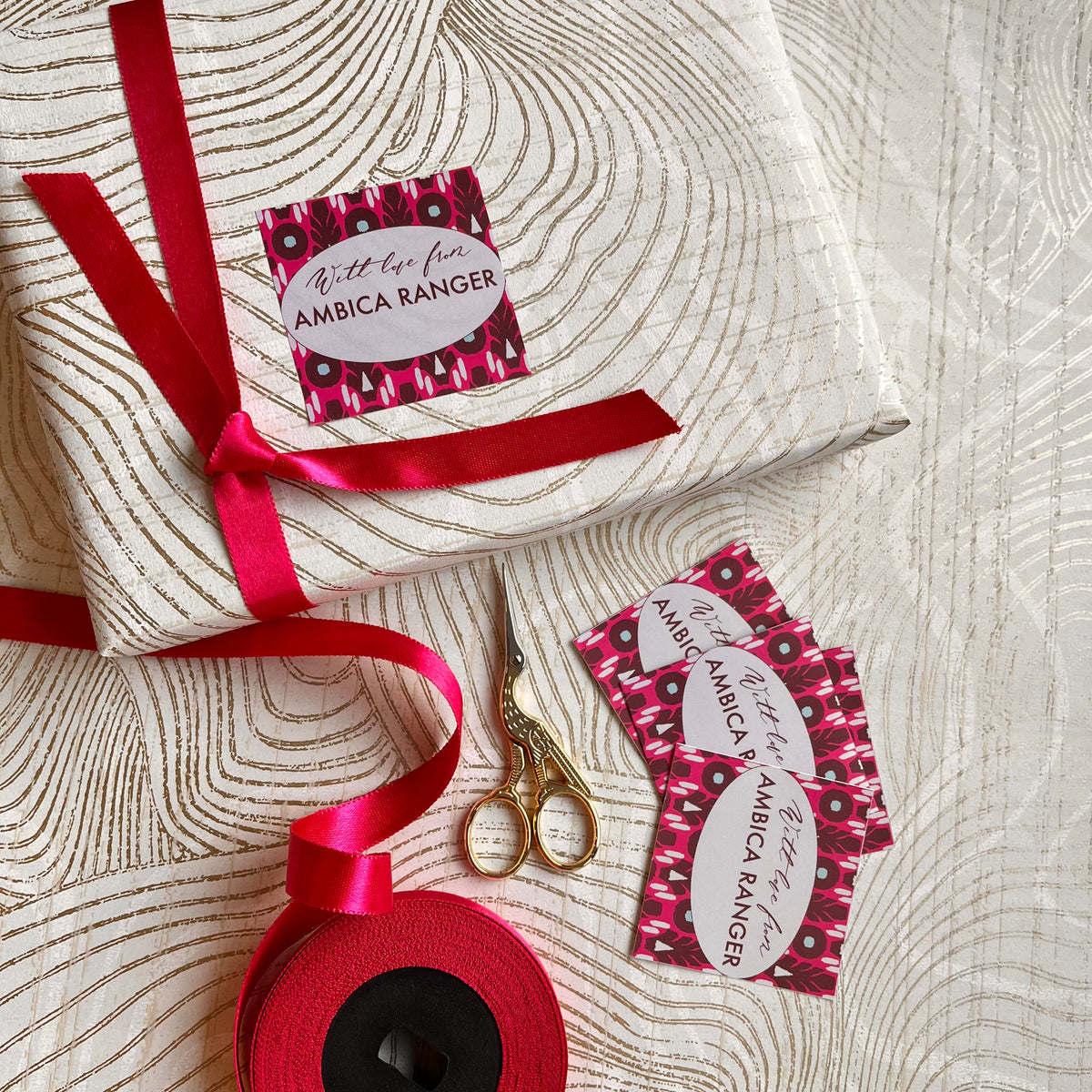 A wrapped gift box tied with a red ribbon, shown with matching red ribbon spool, gold-handled scissors, and several decorative gift tags printed with the Ambica Ranger logo on a light wood-grain surface.