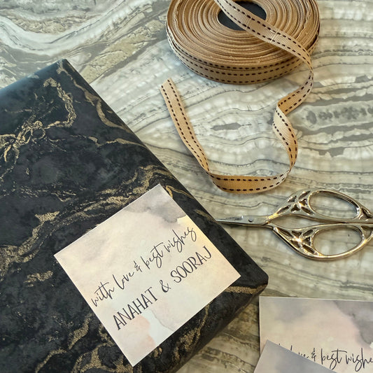 A styled flat-lay of gift wrapping paper featuring a black and gold marble pattern, shown alongside a roll of tan dotted ribbon, silver scissors, and handwritten cards on a gray and white marble-patterned surface.