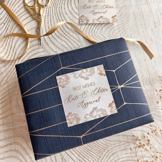 A wrapped gift box in navy blue wrapping paper with a geometric gold line pattern, styled on a light wood grain surface with a gold ribbon, gold scissors, and a small decorative card.
