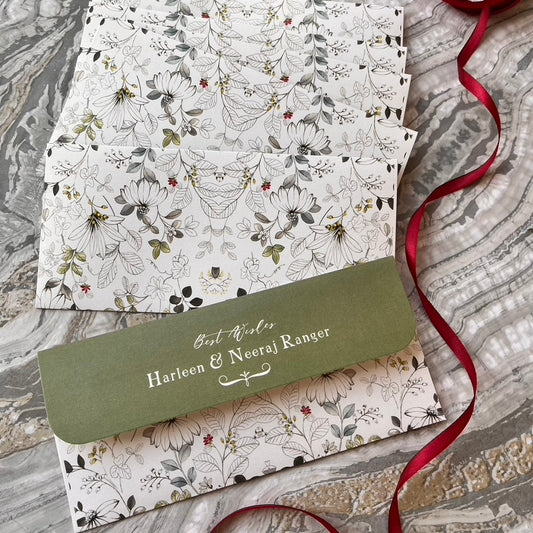A stack of personalized floral gift envelopes with a green flap reading 'Best Wishes Harleen & Neeraj Ranger,' arranged on a gray marble surface with a red ribbon.