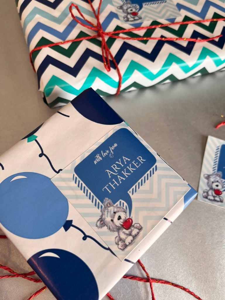 Personalized baby shower gift wrap featuring a blue and white chevron pattern, blue balloon illustrations, and a cartoon teddy bear holding a red heart. The wrap is shown wrapped around a gift box tied with red string, with a matching card visible on the side.