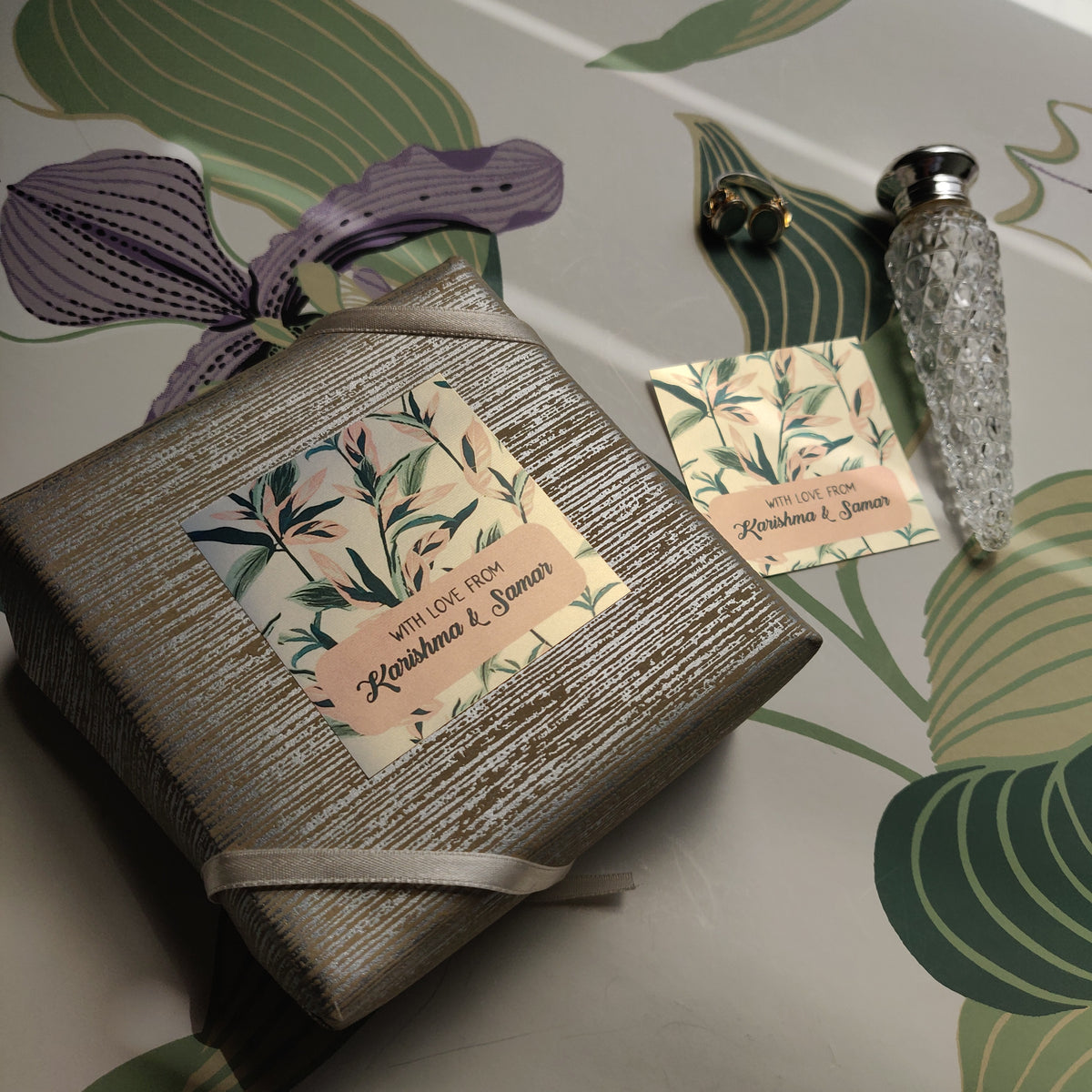 A square wrapped gift box with a textured metallic silver-gray exterior and a floral paper label reading 'A Little Love Always,' styled on a leaf-patterned surface with a small bottle and card nearby.