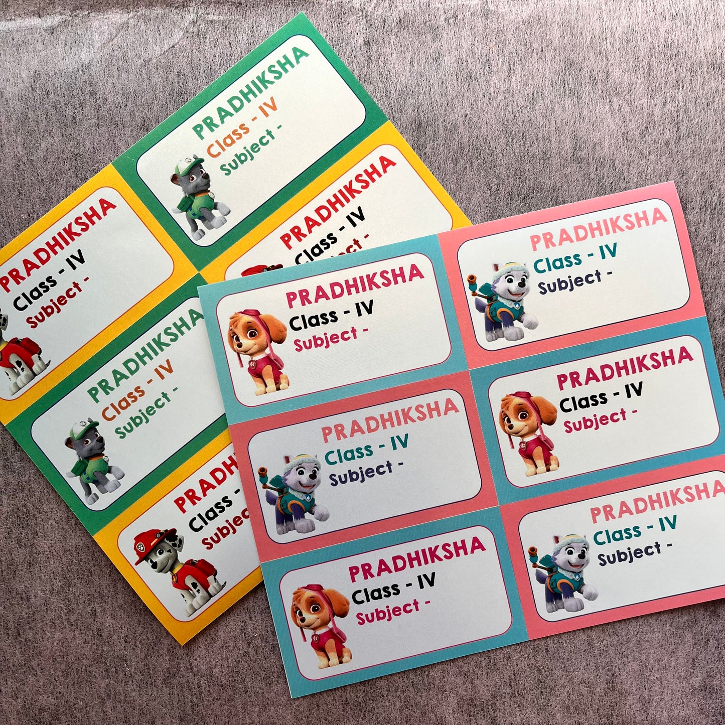 Customizable Paw Patrol-themed name tags arranged on a textured gray fabric background. The tags feature various character designs (Puppy, Chase, Rubble, Skye, Marshall, Zuma) with different border colors including green, yellow, orange, pink, and blue.
