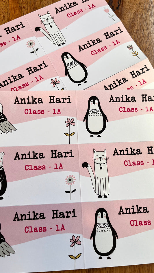 personalised printed book name labels with pink and white sections, black text reading 'Anika Hari Class - 1A', and cute cartoon illustrations of a cat, penguin, and flower,