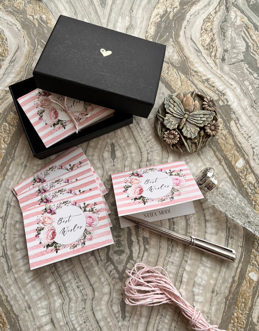 A styled flat-lay of a bridal shower favor kit on a marble-patterned surface. The kit includes a black gift box with a small gold heart, pink and white striped envelopes with floral designs and 'Best Wishes' writing, a silver fountain pen, pink thread, and a decorative butterfly ornament.