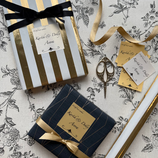 Two wrapped gift boxes on a floral-patterned surface, one with white and gold stripes and a black ribbon, and one with dark navy and gold stripes and a gold ribbon. A gold tag, scissors, and ribbon are arranged around them.